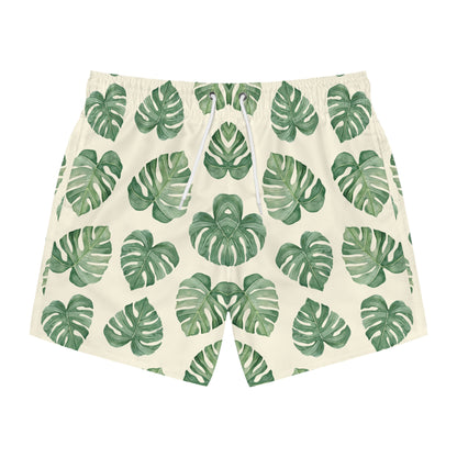 A pair of men's swim shorts featuring an all-over green monstera leaf pattern on a warm cream background, with a white drawstring at the elastic waistband.