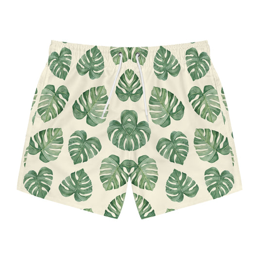 A pair of men's swim shorts featuring an all-over green monstera leaf pattern on a warm cream background, with a white drawstring at the elastic waistband.