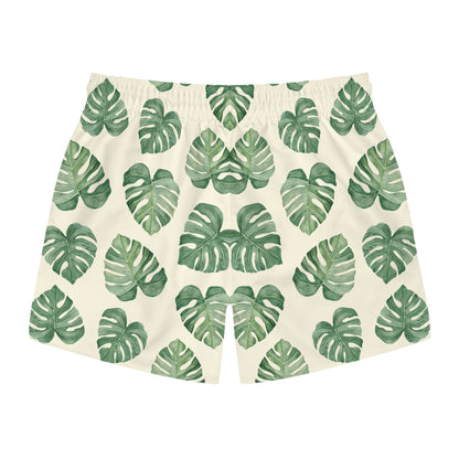 A pair of men's swimming trunks  featuring an all-over green monstera leaf pattern on a warm cream background, displayed flat against a solid white background.